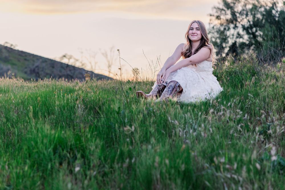 A person in a white dress sits peacefully in tall grass on a hillside during golden hour sunset.