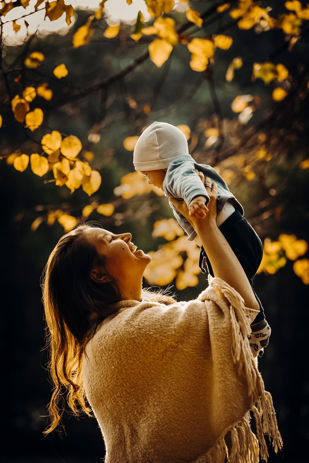 Persona con un maglione comodo che tiene in braccio un bambino sullo sfondo di un fogliame autunnale dorato.