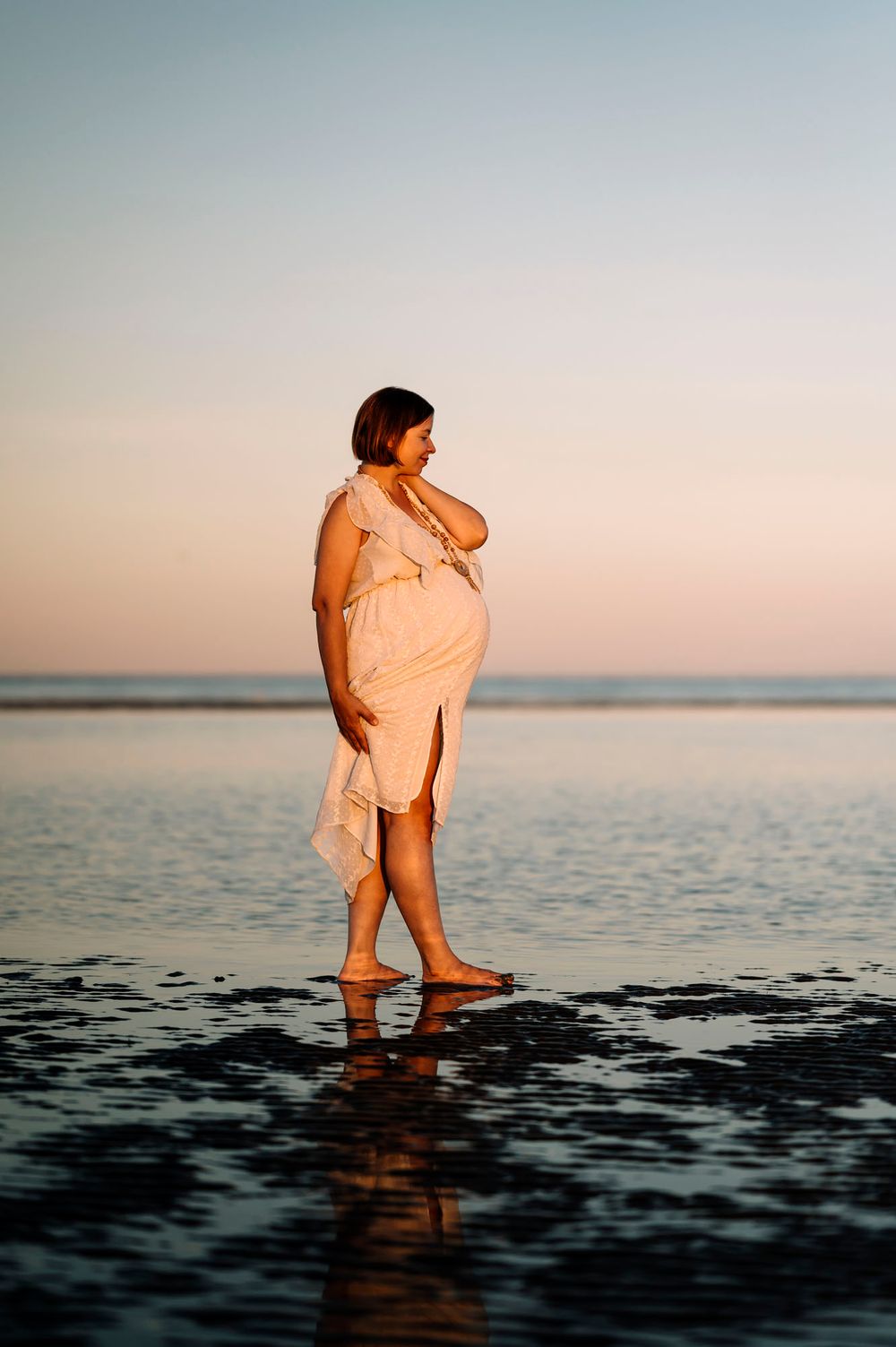 Una donna in dolce attesa con un abito rosa svolazzante cammina nell'acqua bassa al tramonto creando riflessi sulla sabbia scura e bagnata