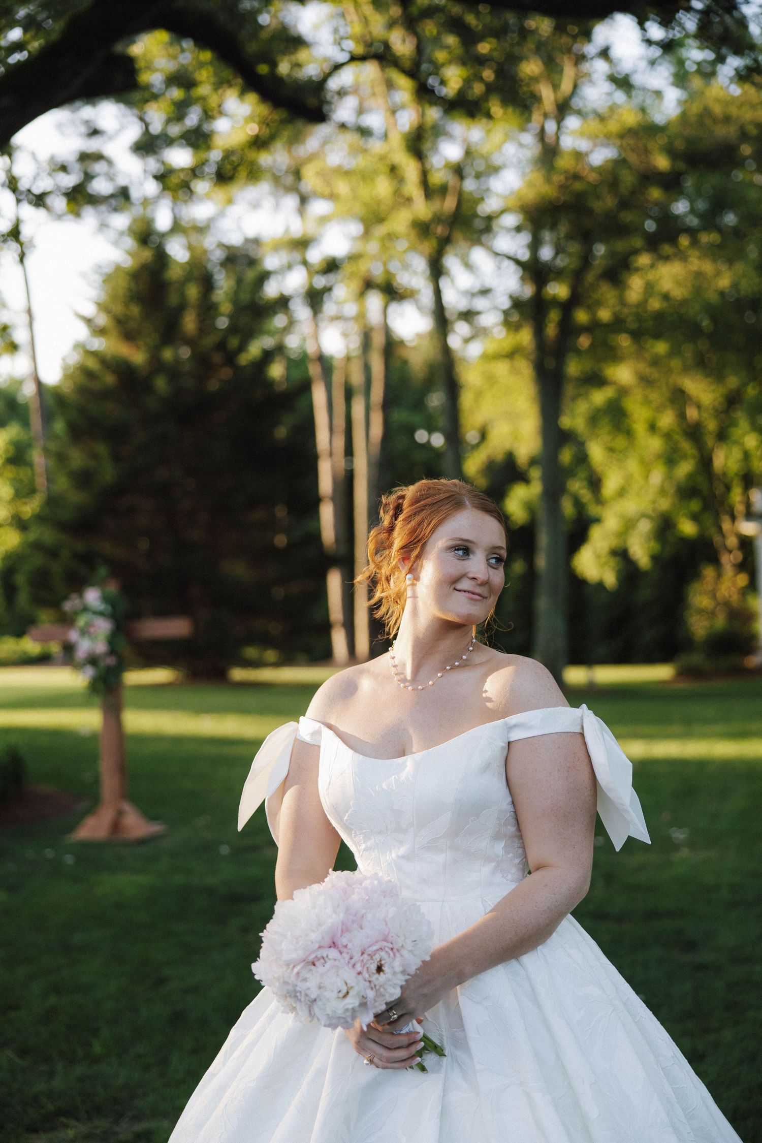A bride in a white off-shoulder ball gown holds a pink bouquet while standing in a sunlit garden setting.