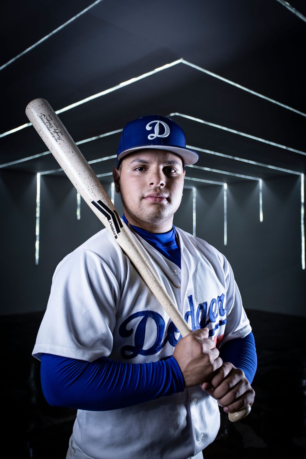 Baseball player in Los Angeles Dodgers uniform poses in dramatic lighting against dark background with batting stance.