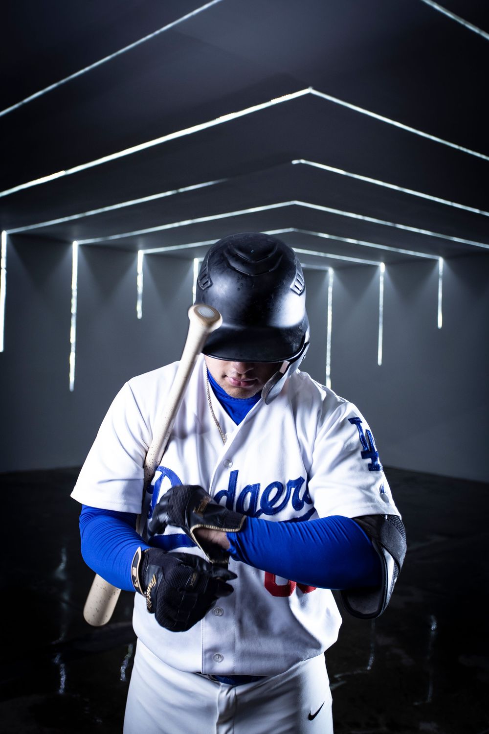 Baseball player in white Dodgers jersey and helmet looking down at his hands in dramatic lighting.