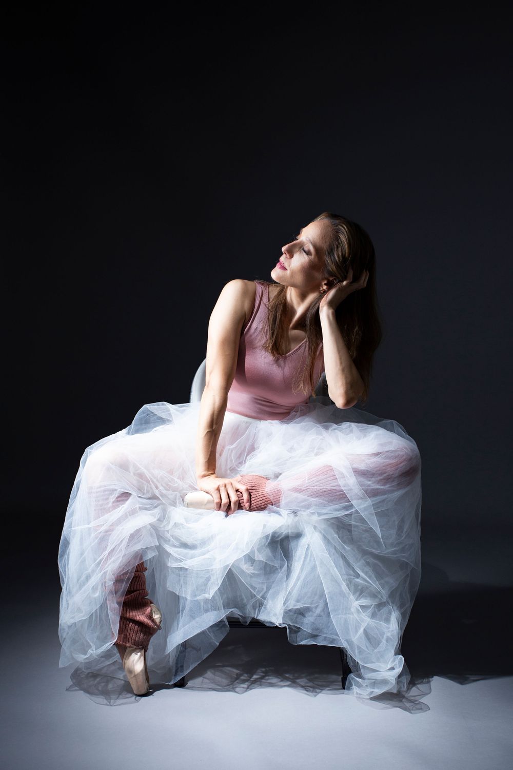 Graceful ballet dancer in pink leotard and white tulle skirt poses elegantly in dramatic studio lighting.