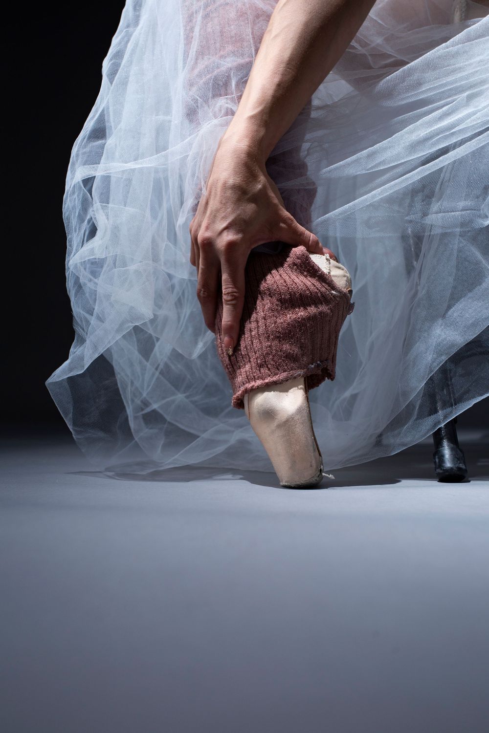 A ballet dancer's feet in pointe shoes with flowing white tulle fabric and a warm knit leg warmer.