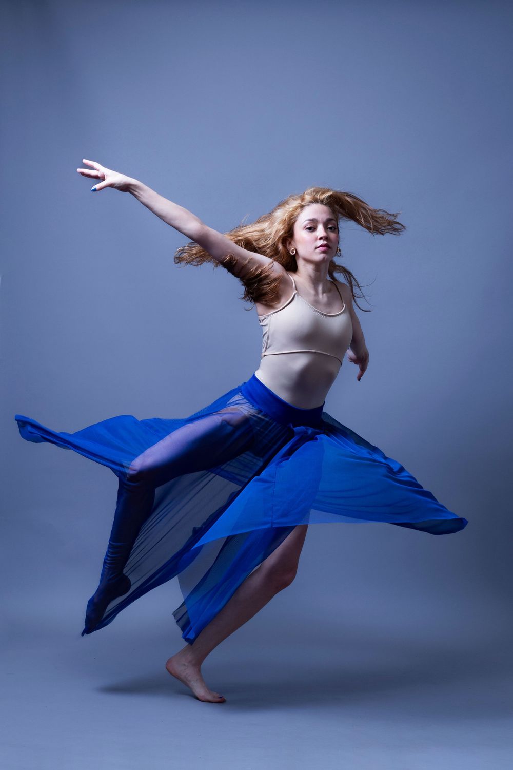 A dancer in a white top and flowing blue skirt performs an expressive modern dance movement against a blue backdrop.
