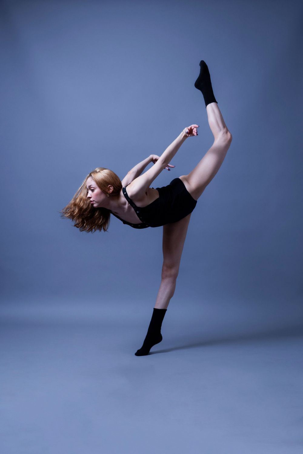 Modern dancer in black leotard performing dynamic kicks and movements against blue studio backdrop.