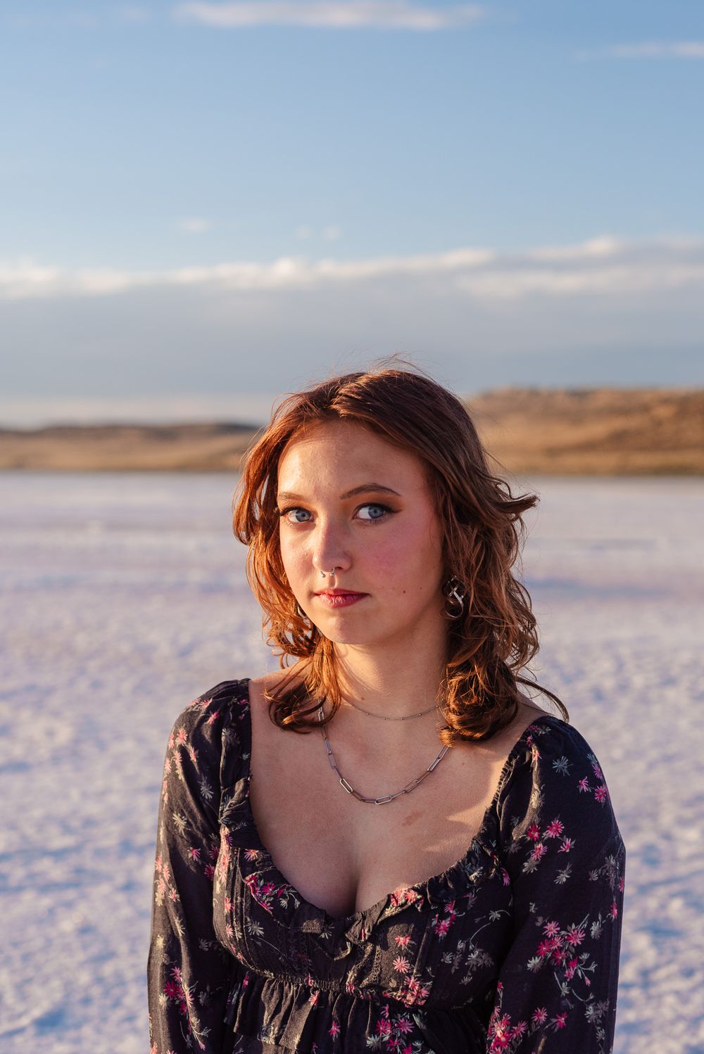 Red-haired teenage girl in floral dress with necklace posing on bright white salt flats under blue sky.