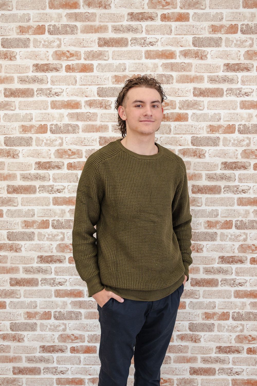 Young man in olive green knit sweater standing casually with hands in pockets against a brick wall.