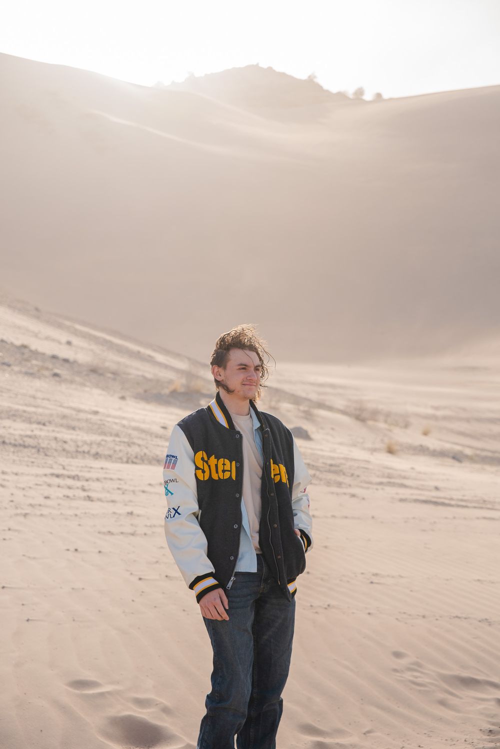 Young man in Steelers varsity jacket standing on sandy desert dunes.