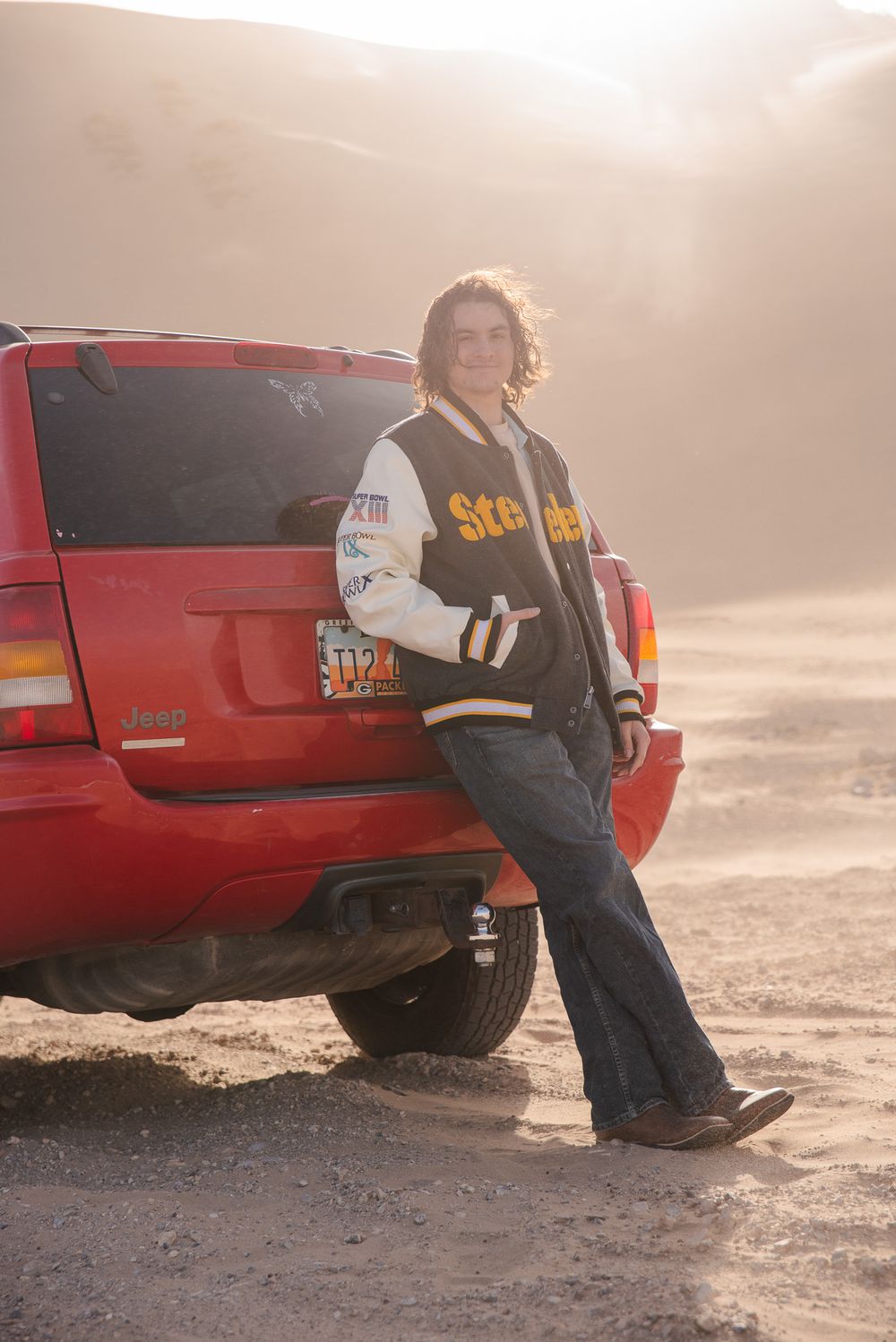 Young man in Steelers varsity jacket posing beside red SUV in desert.
