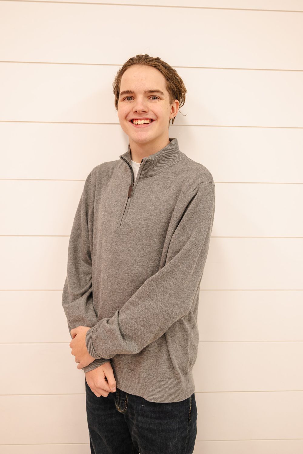 Smiling teenage boy with brown hair wearing a gray quarter-zip pullover against a white shiplap wall.