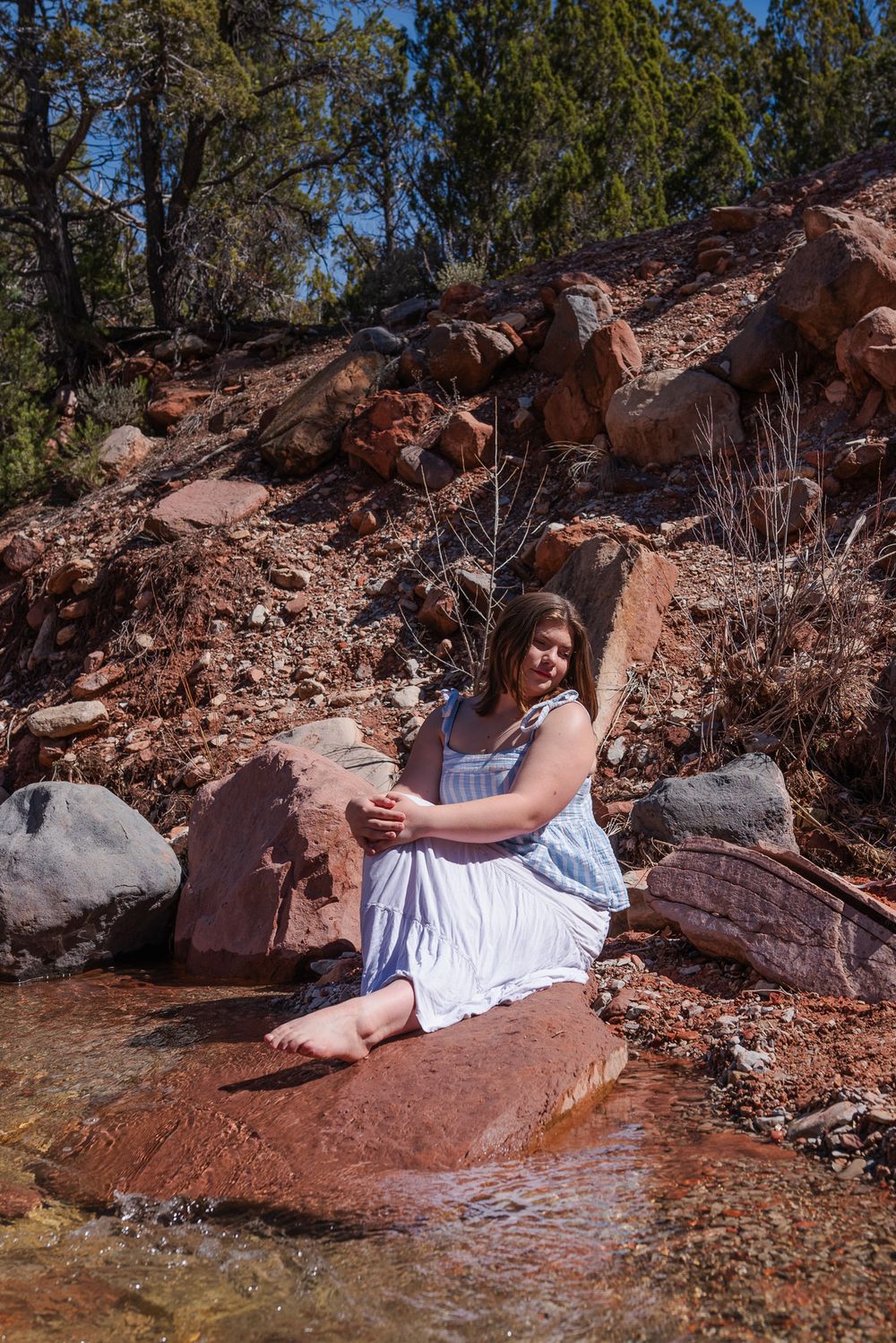 Young woman in blue striped dress sits barefoot on red rocks beside a stream in a desert canyon.