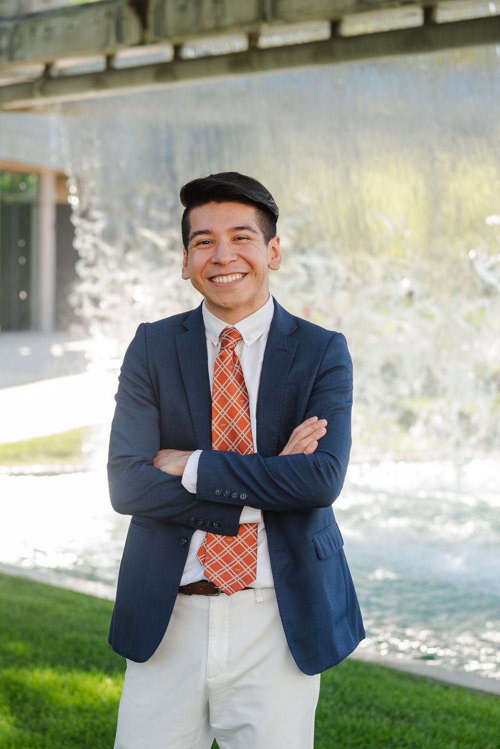 Smiling young man in navy suit and orange tie standing outdoors with arms crossed.