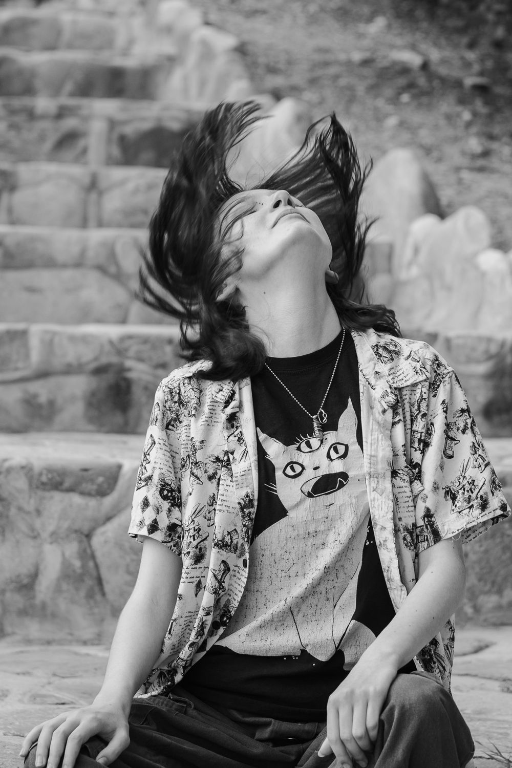 Black and white photo of a person flipping long hair back while sitting on stone steps outdoors.