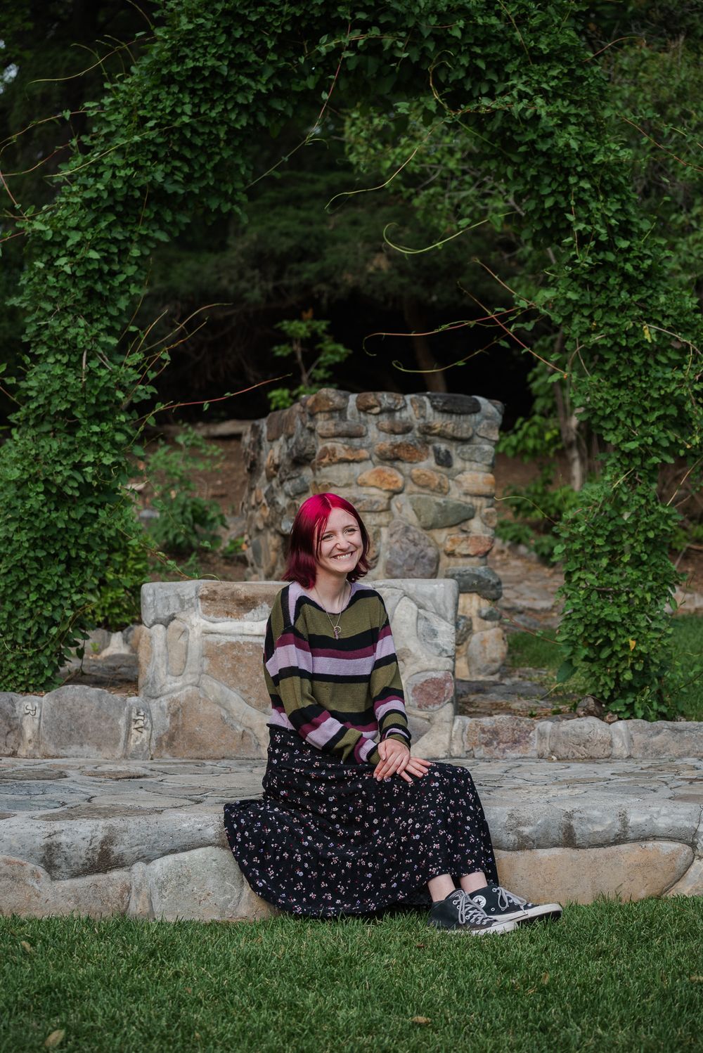 Smiling young woman with pink hair in a striped sweater and floral skirt sitting on stone steps outdoors.