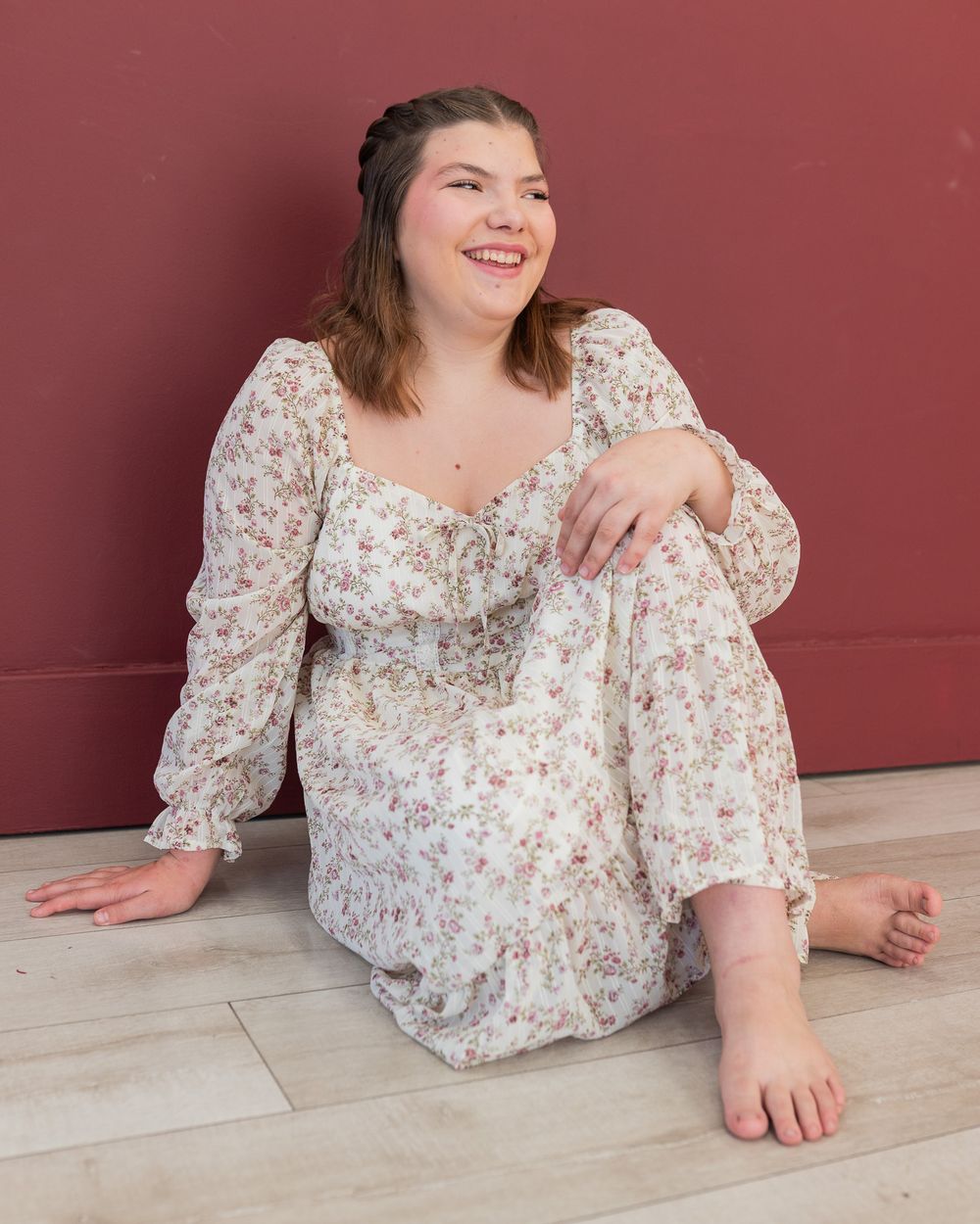 Young woman in floral dress sitting barefoot on floor smiling against a red wall.