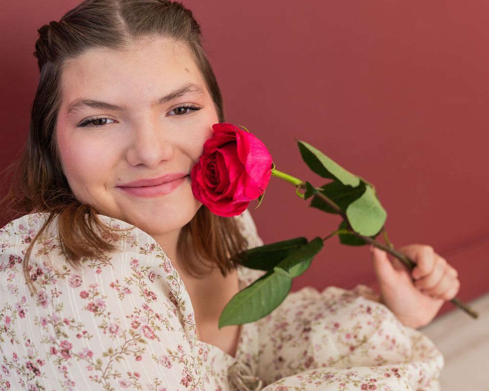 Young woman smiling while holding a red rose close to her cheek indoors.