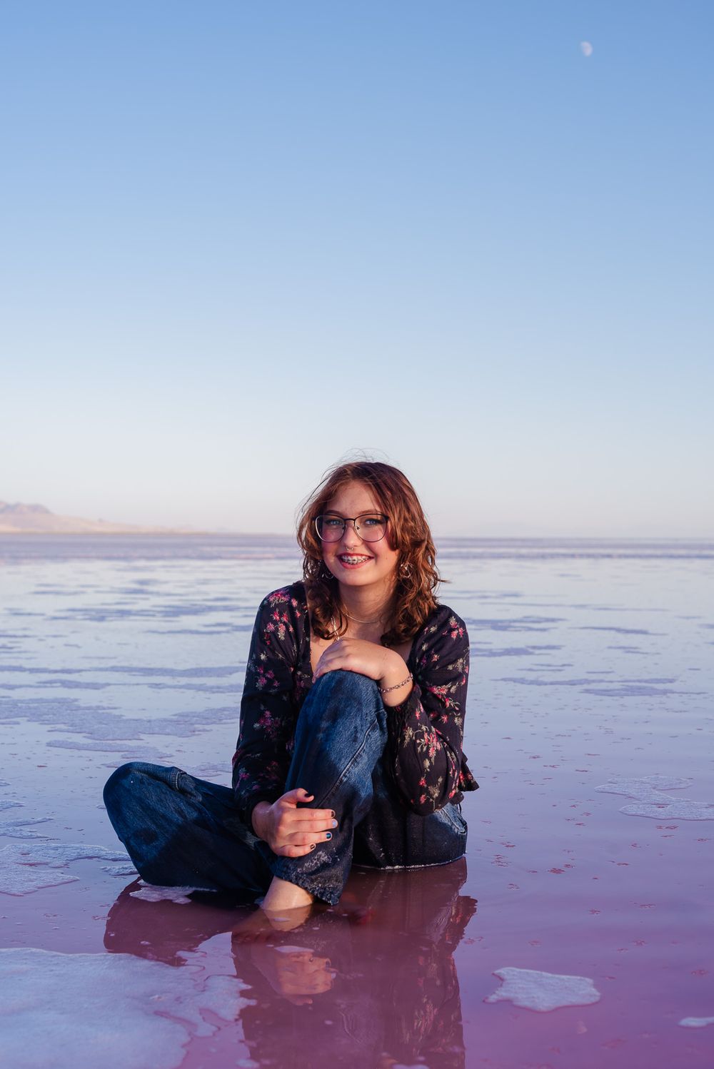 Smiling woman with glasses sitting cross-legged on pink salt flats at dusk with moon visible.