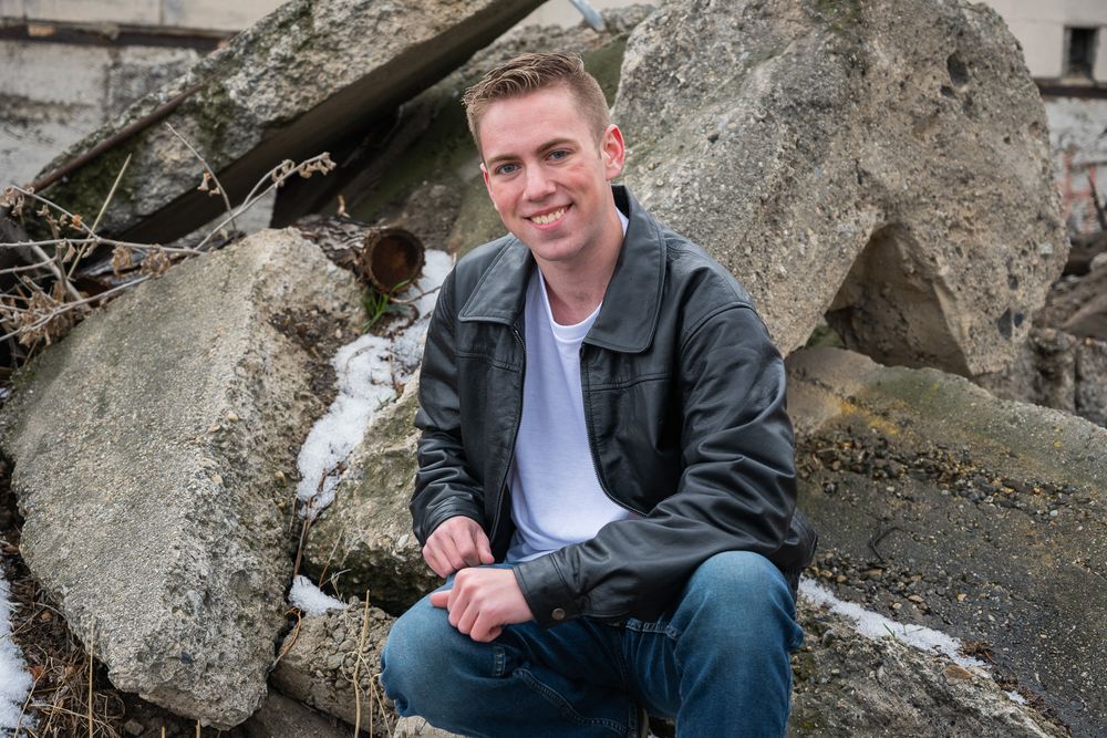 Smiling young man in black leather jacket sitting on rocks outdoors in winter.