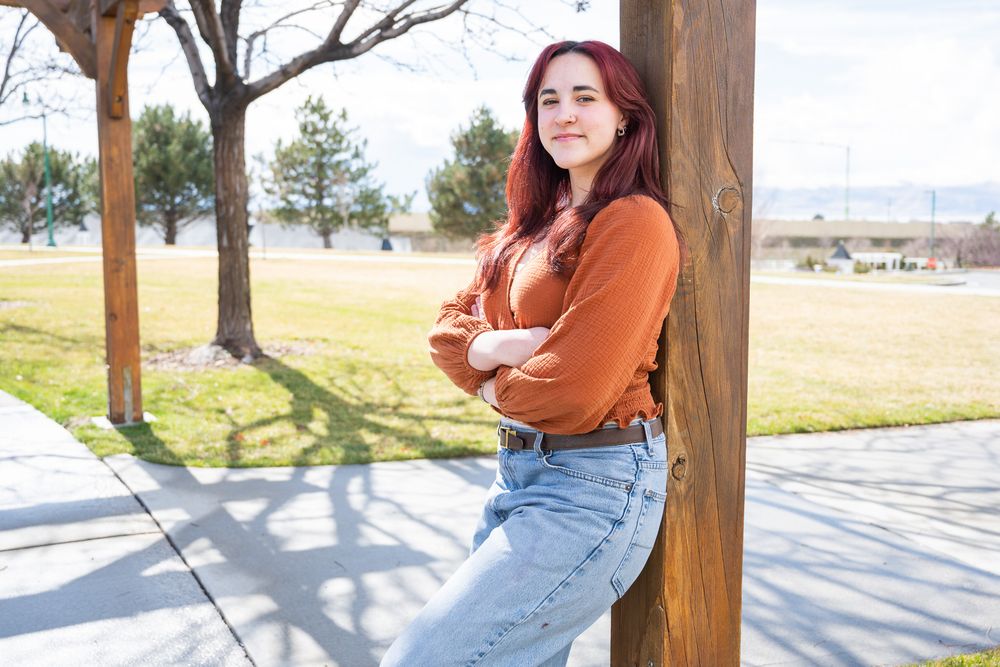 Young woman with red hair in orange sweater leaning against wooden post outdoors.
