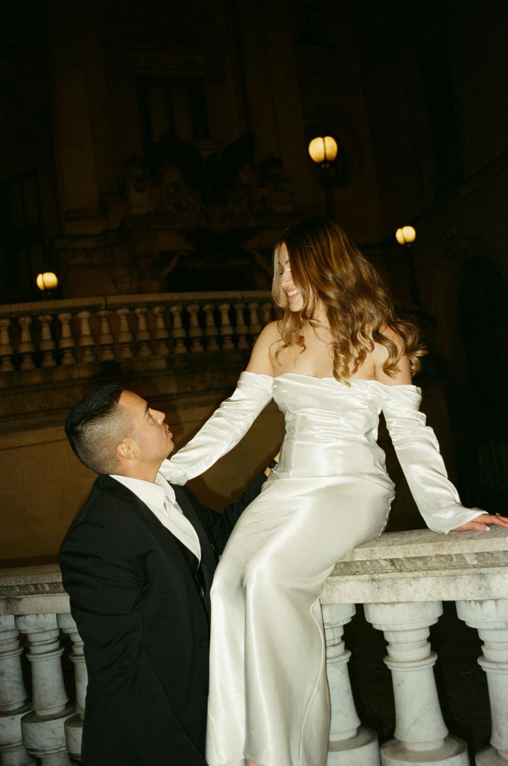 A Paris couple in wedding attire share a romantic moment near an ornate stone railing in a dimly lit venue. film photography.