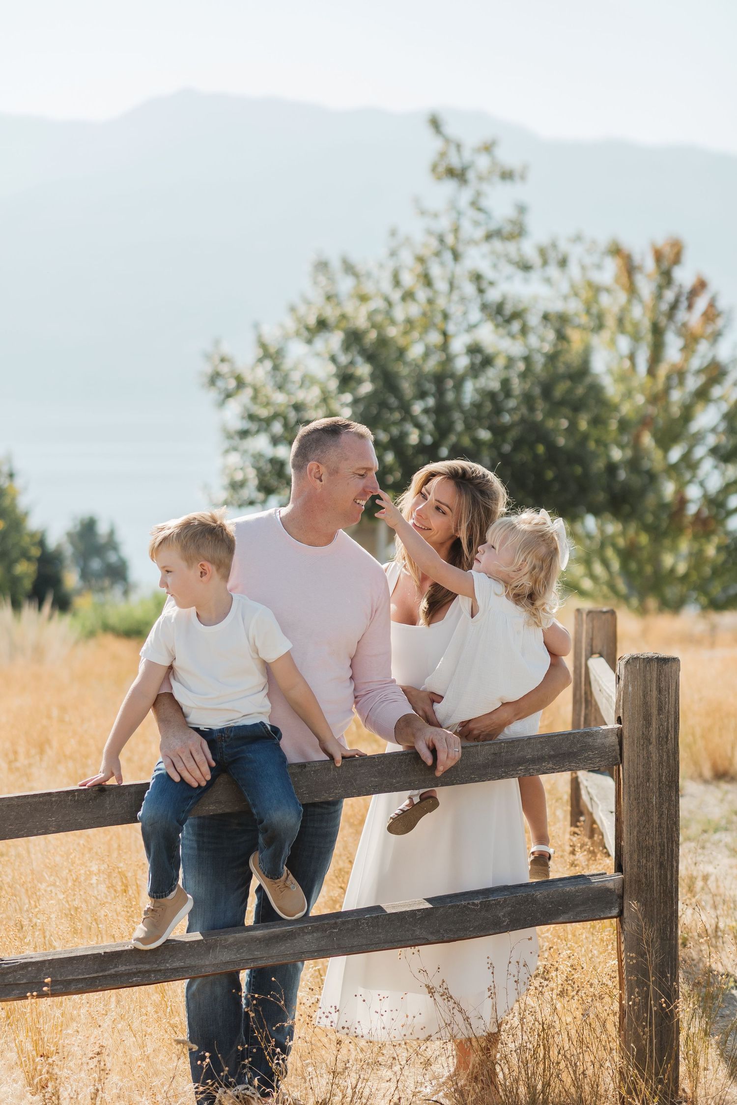 Family in white and denim outfits sitting and laughing together on rustic wooden fence in golden field at sunset.
