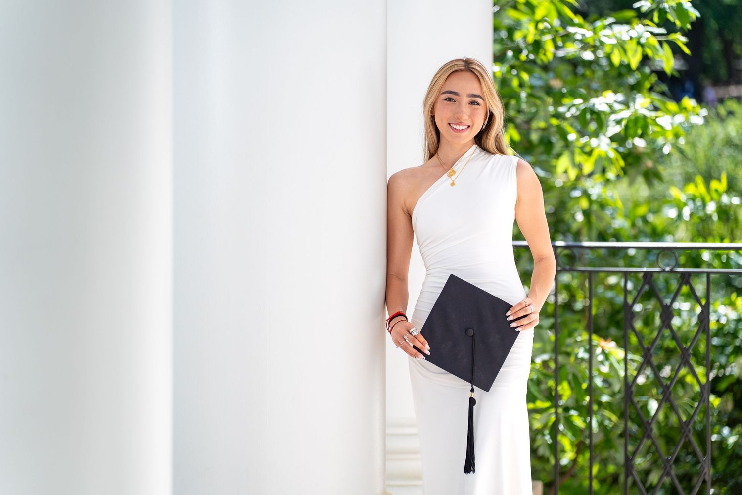 Graduate in white dress poses joyfully against column while holding graduation cap.