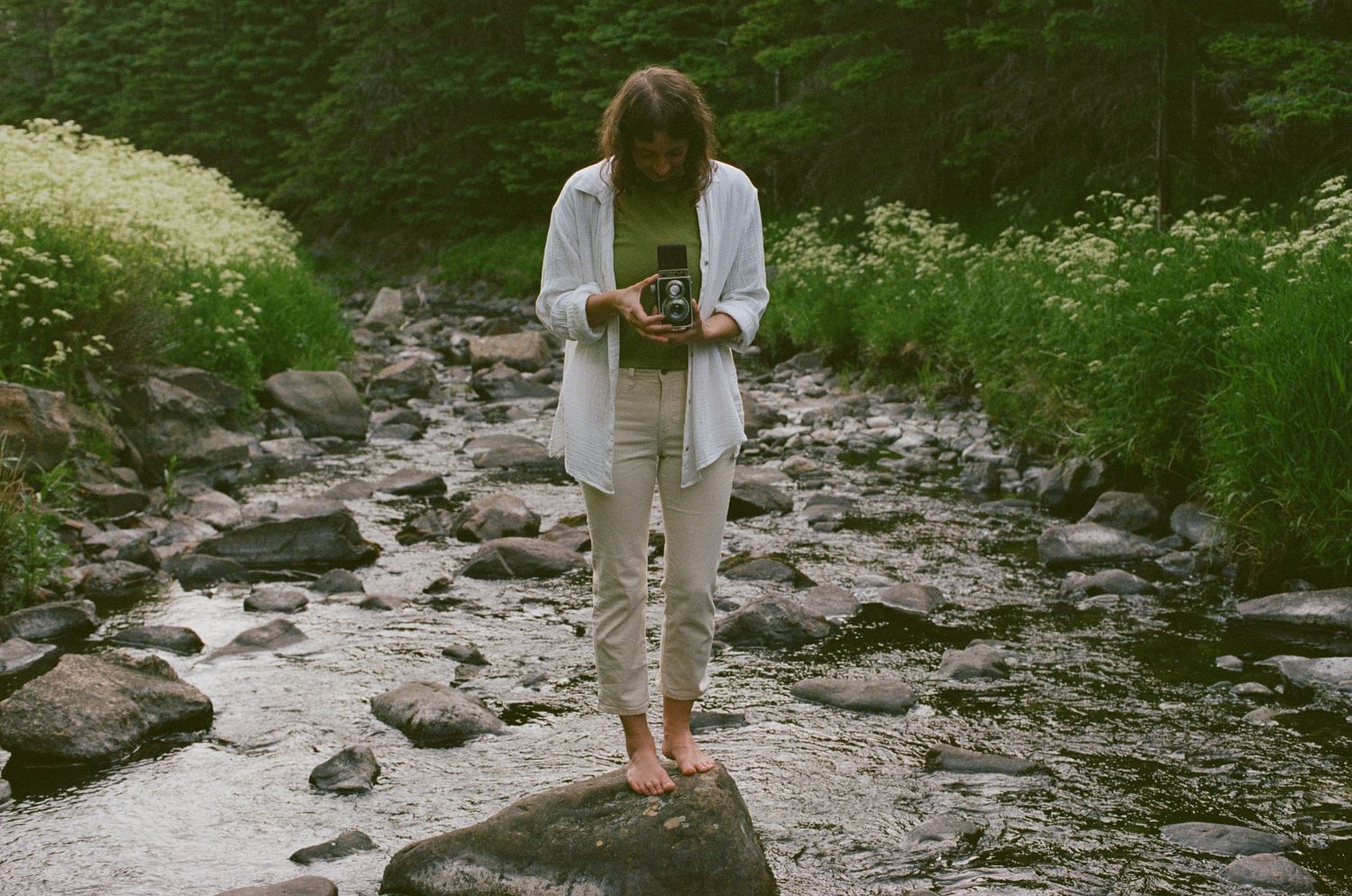 Heather Nolan of Wildflower Elopement Co stands on a rock in a river surrounded by wildflowers, looking down into a TLR film camera