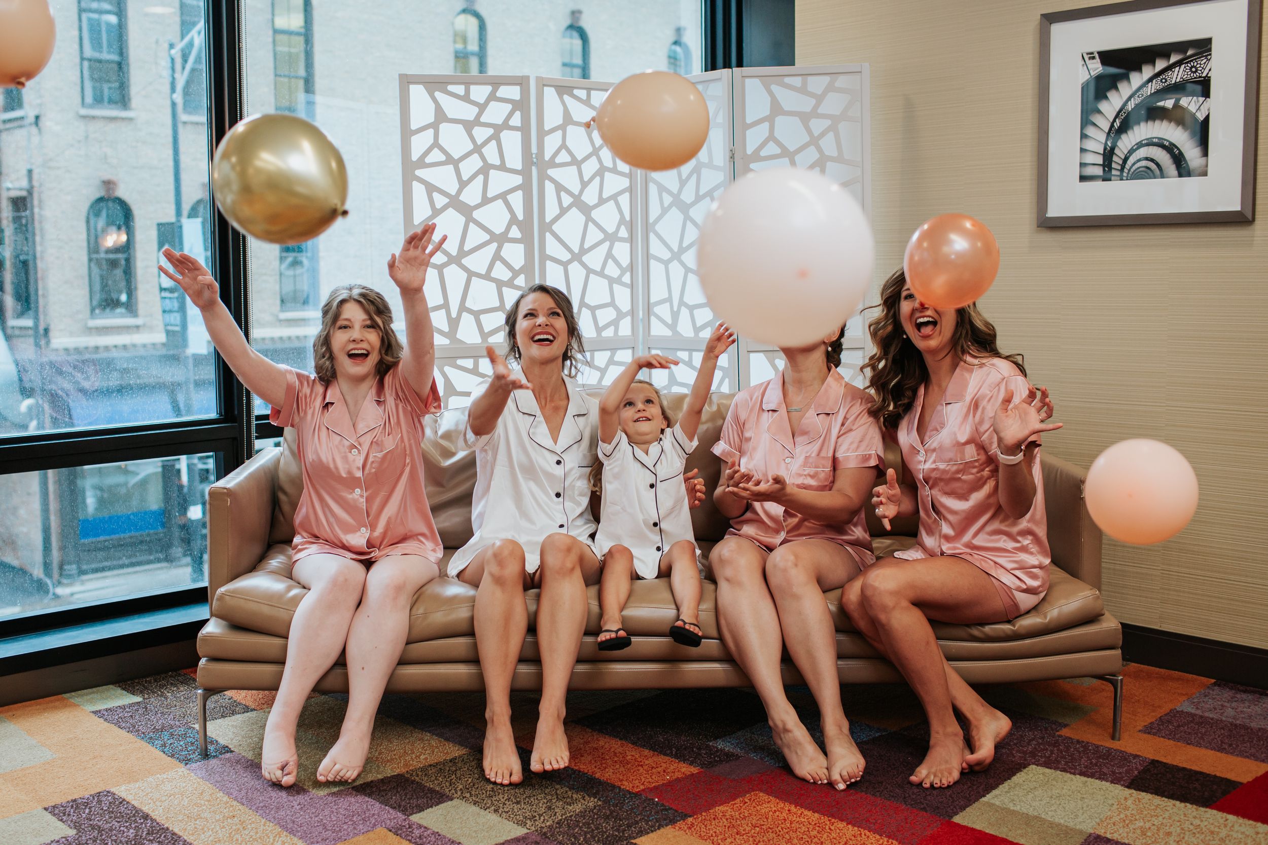 Friends in matching pink robes sit together on a couch with balloons during a celebration.
