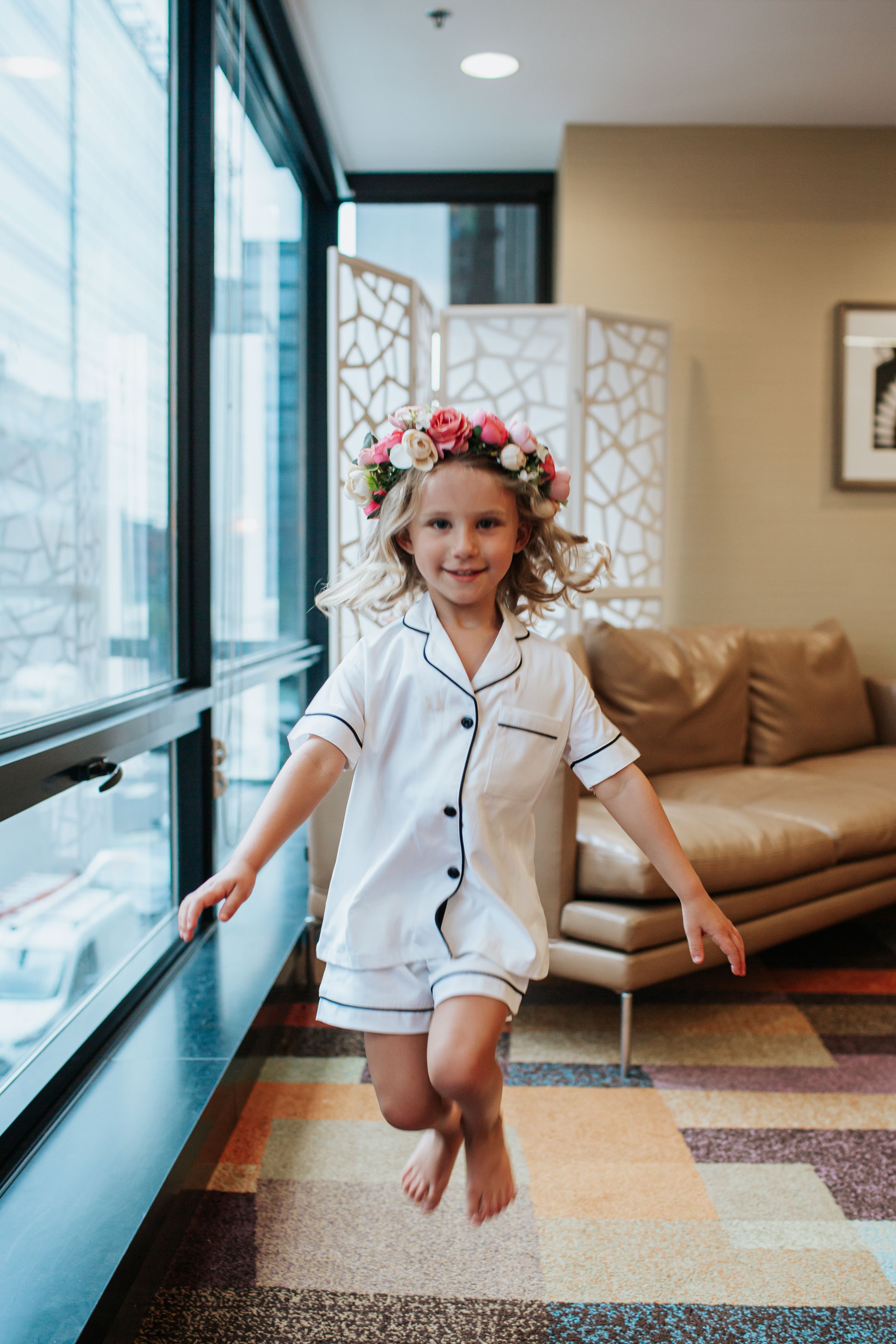 Child in white pajamas jumping joyfully by window wearing floral crown.