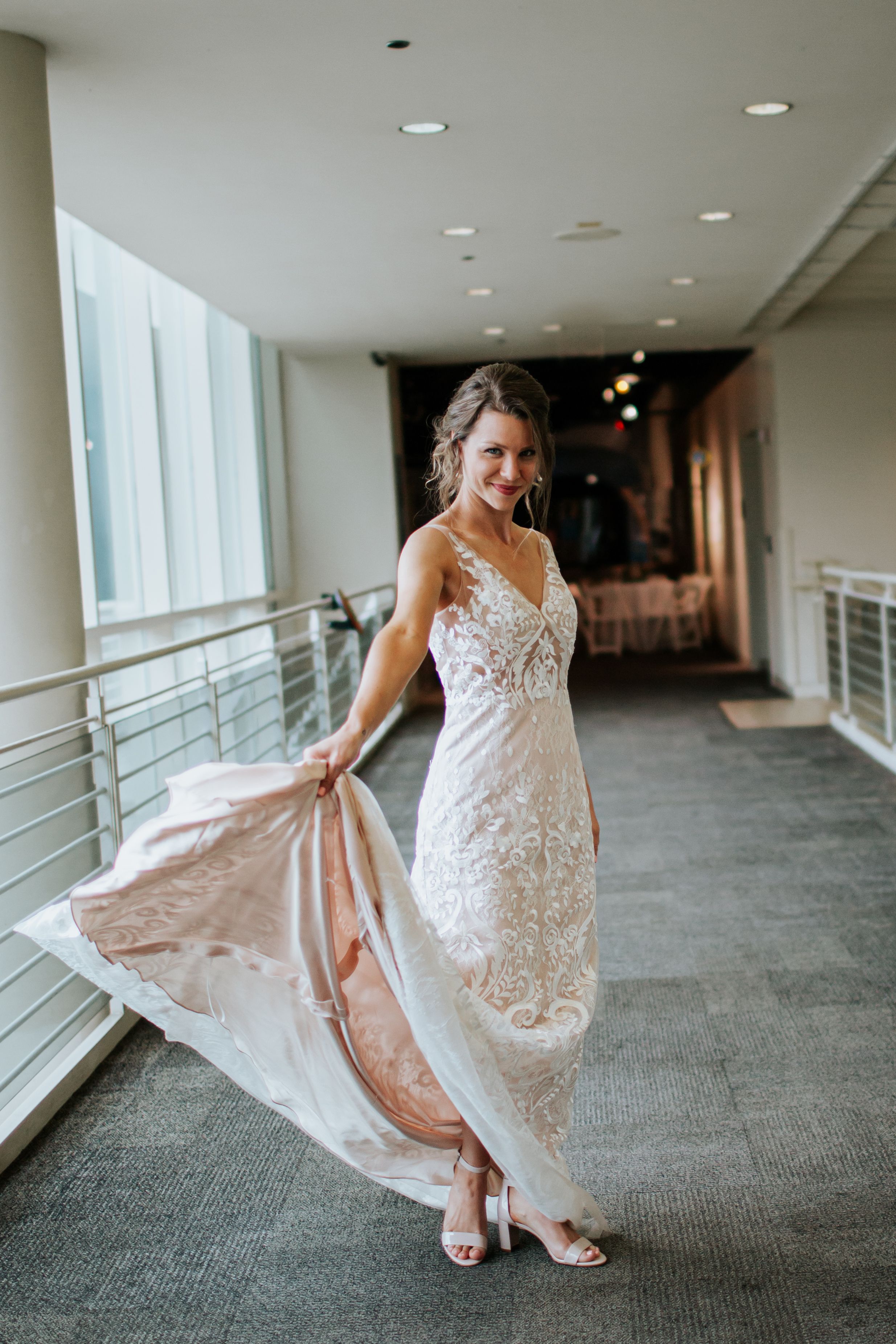 A person wearing a white lace wedding dress twirls playfully in a modern hallway with floor-to-ceiling windows.