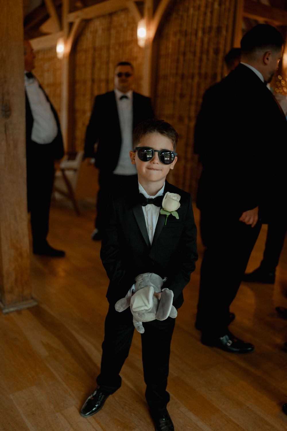 A young ring bearer in a black tuxedo and sunglasses smiles while holding a stuffed animal at a wedding.