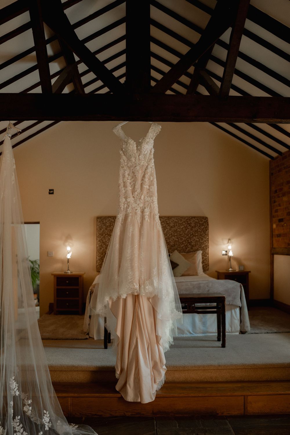 Elegant lace wedding dress hanging in a warmly lit rustic bedroom with wooden beam ceiling and soft ambient lighting.