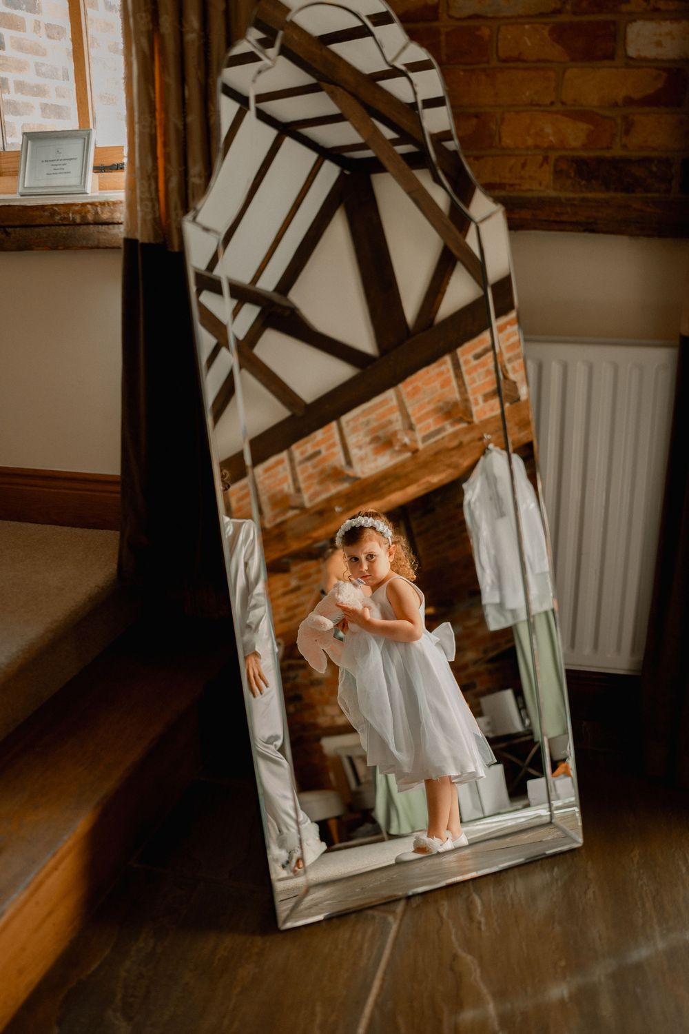 Young flower girl in white dress reflected in ornate mirror inside rustic barn wedding venue.