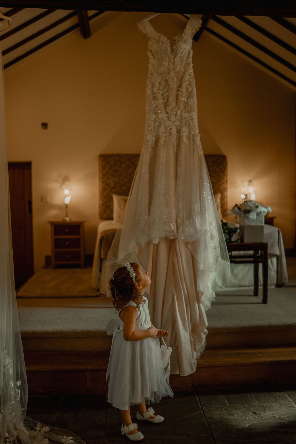 A young flower girl in white dress gazes up at an elegant lace wedding gown hanging in a warmly lit bridal suite.
