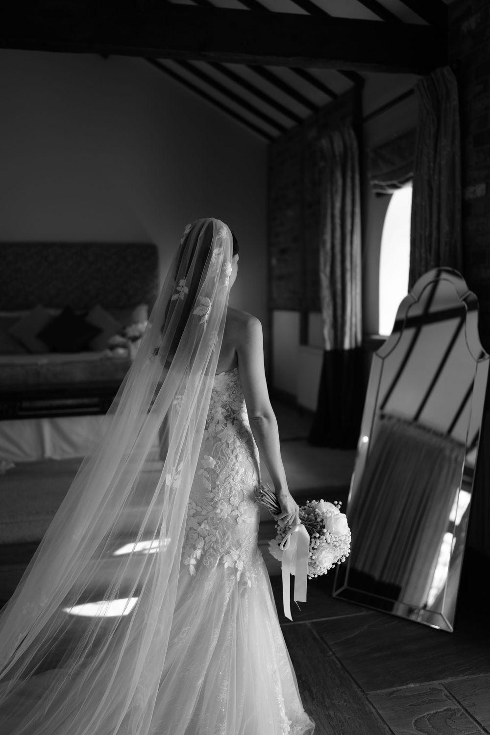 Black and white photo of a bride in a flowing gown and veil holding a bouquet, standing before a mirror in an elegant room.