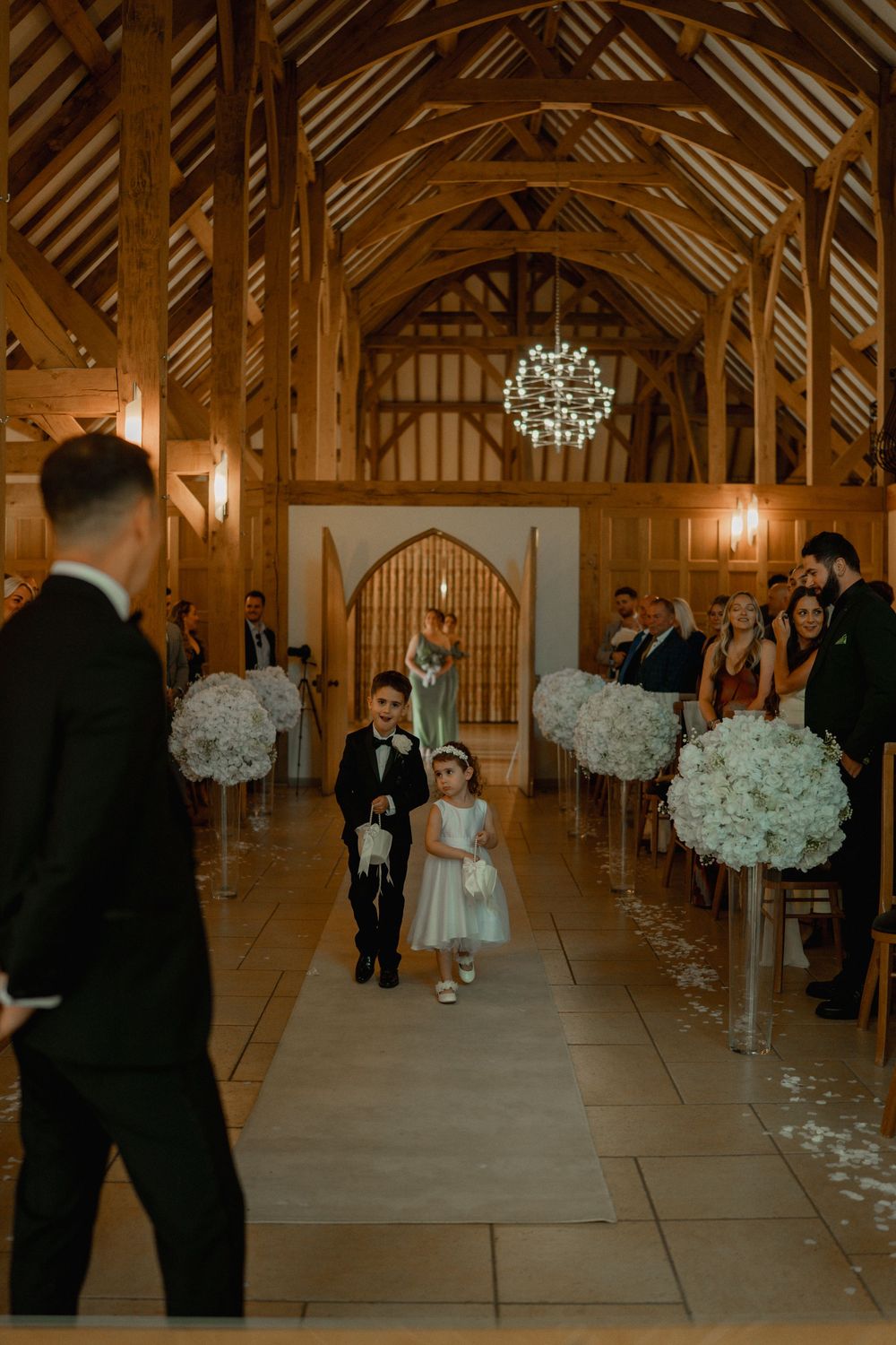 A flower girl and ring bearer walk down the aisle in an elegant wooden barn wedding venue with white floral arrangements.