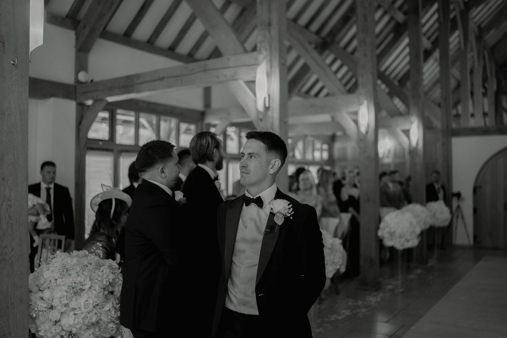 Groom in tuxedo and bow tie stands in elegant barn wedding venue, black and white photography.