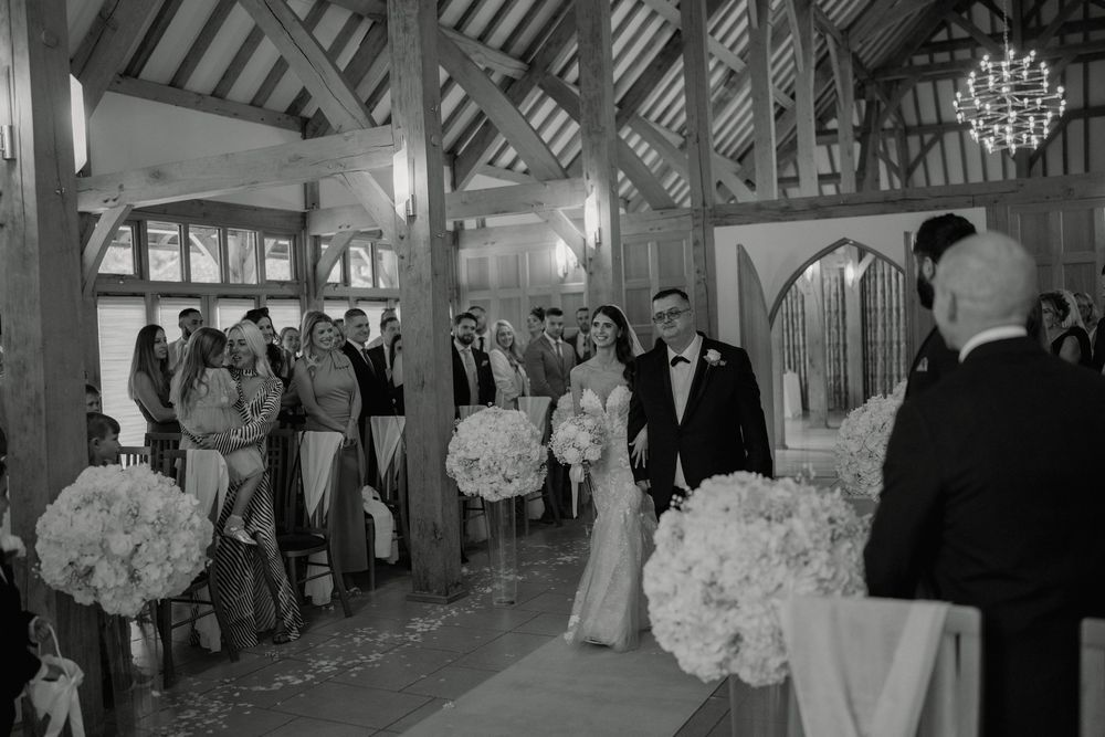 Black and white wedding ceremony photo of bride and groom walking down the aisle in a rustic barn venue.