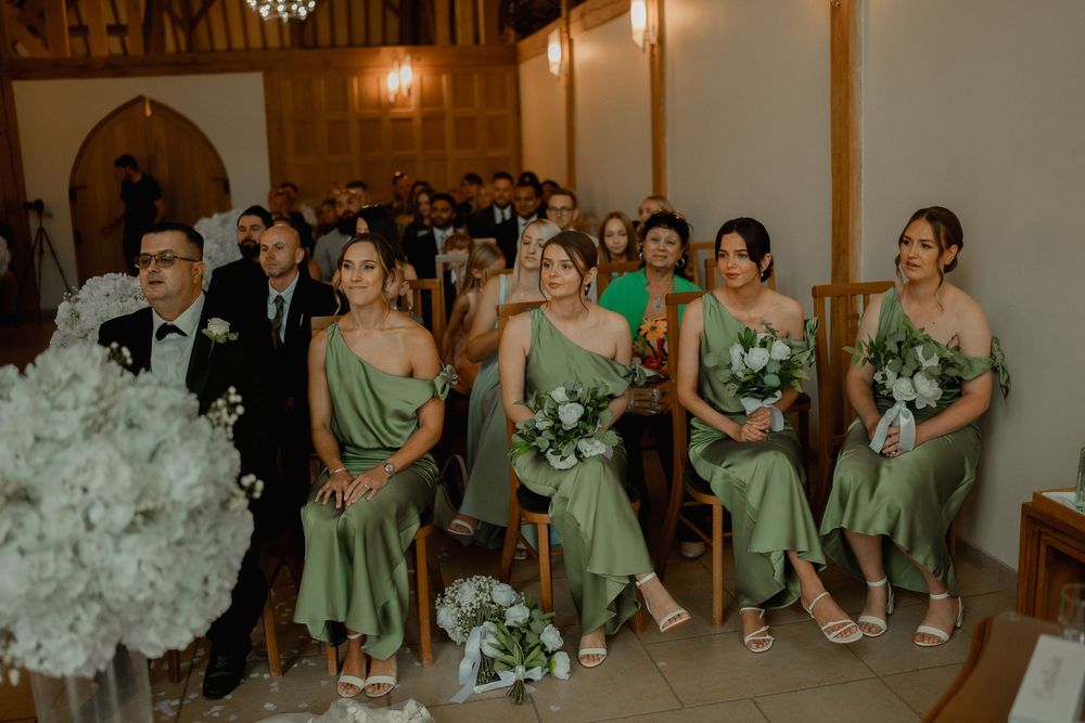 Bridesmaids in sage green satin dresses holding white bouquets seated in a rustic barn wedding ceremony.