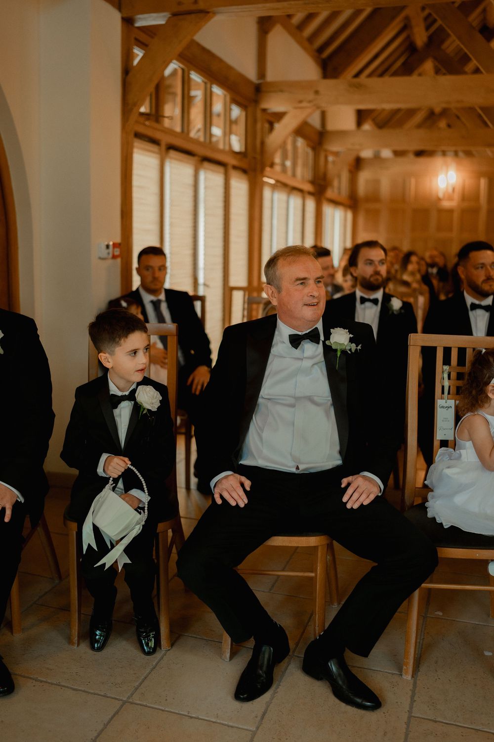 A man and young boy in matching tuxedos sit together at a wedding ceremony in an elegant wooden venue.