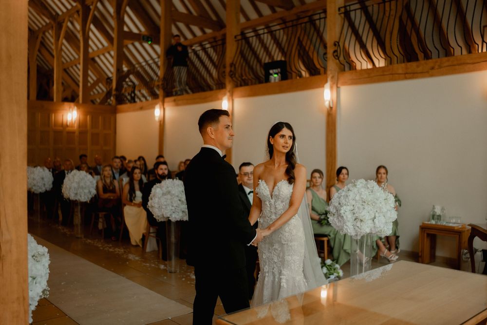 Bride and groom hold hands at rustic barn wedding ceremony with white floral arrangements and seated guests in background.