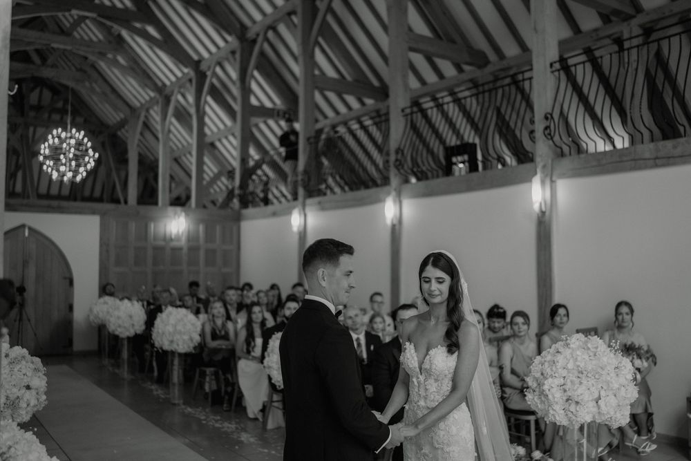 Black and white photo of a bride and groom exchanging vows in a rustic barn wedding ceremony with guests seated.
