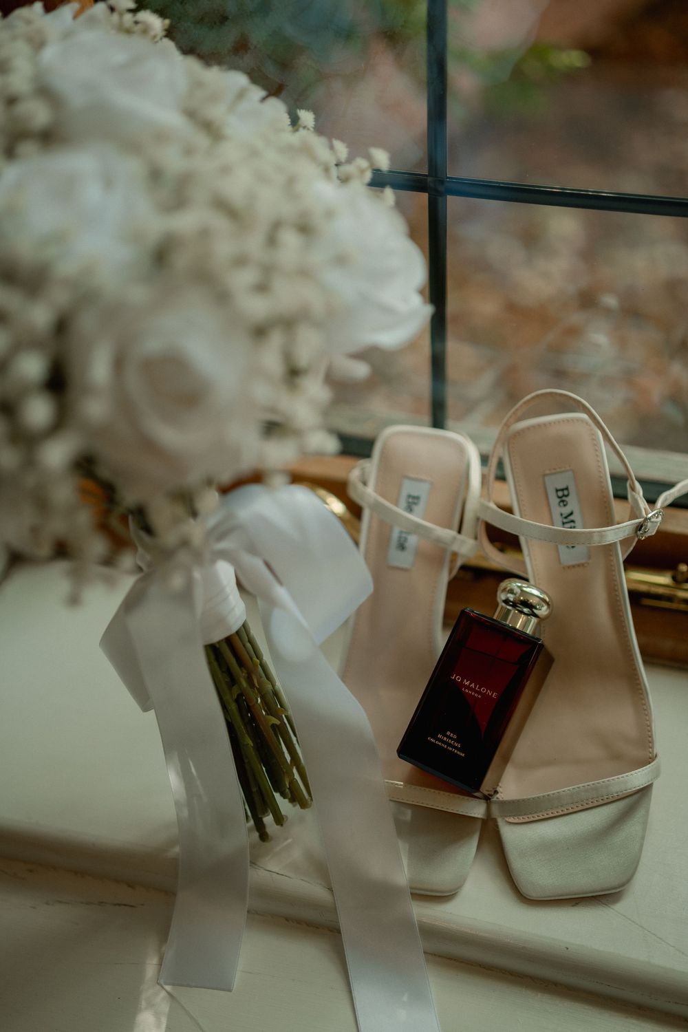 Elegant bridal flat lay featuring white heels, a lush white floral bouquet with ribbon, and a luxury perfume bottle on a windowsill.