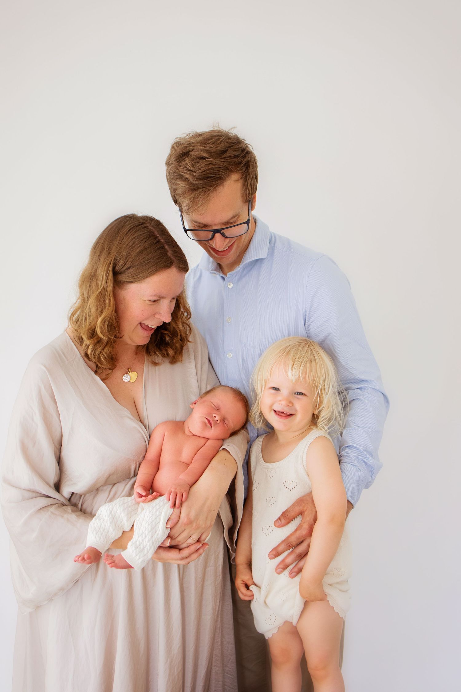 En familj med ett nyfött barn och ett litet barn tillsammans i vita kläder mot en ljus bakgrund för porträtt i fotostudio i Ängelholm