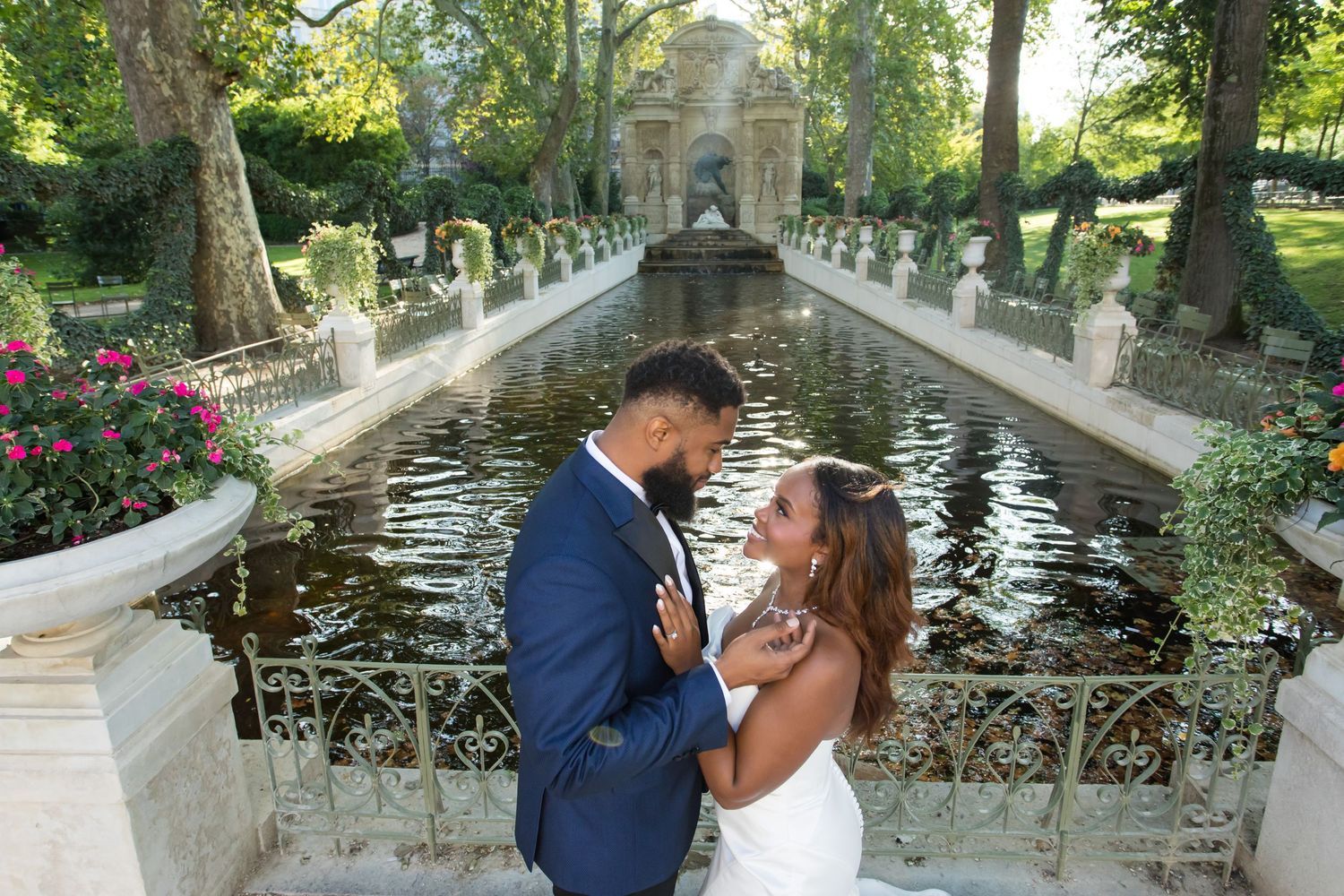 A romantic couple embraces near a tranquil garden fountain of Medicis surrounded by lush greenery and stone architecture.