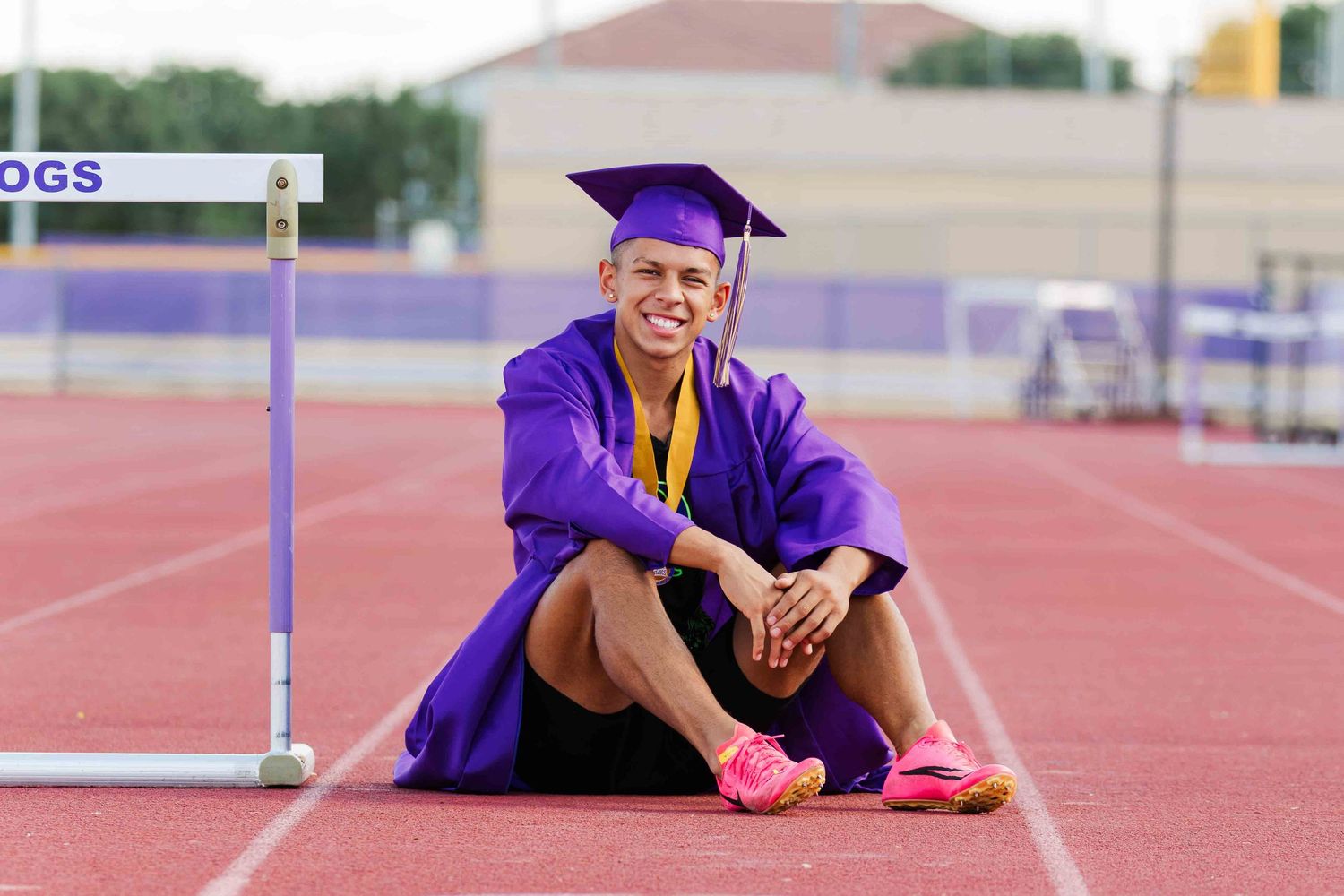 Graduate in purple cap and gown sits on red running track near hurdle wearing pink running shoes.