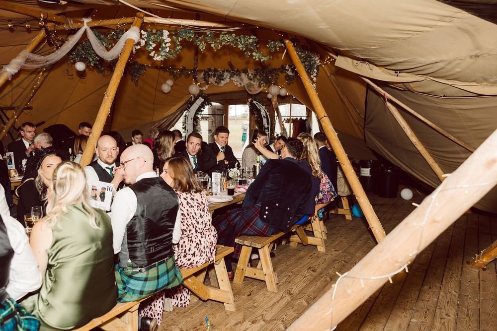 Wedding guests seated at long wooden tables inside a rustic tent decorated with string lights and floral garlands.