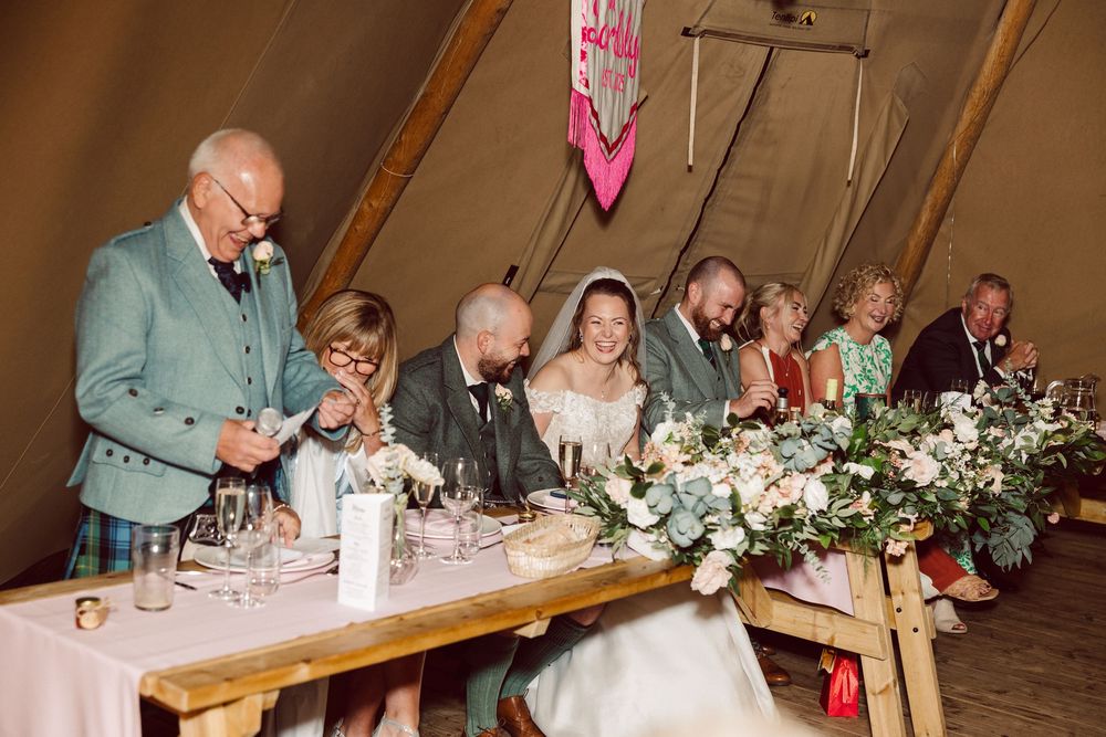 Wedding guests enjoy dinner at a rustic wooden table decorated with white floral centerpieces and candles.