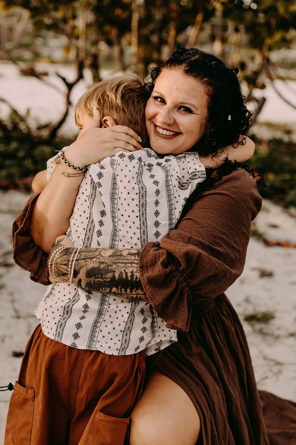 Two people in brown and white dresses embrace warmly in a snowy outdoor setting during a winter photoshoot.